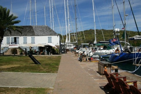 Yachts are moored stern to along the quay in English Harbor. The building to the left is one of several historic structures preserved and now occupied by an internet cafe and a boat electronics service company (note Raymarine banner on steps).